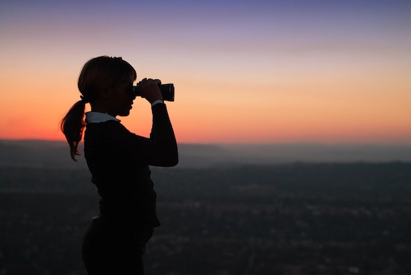 A girl standing on a hill looking through binoculars with a sunset behind her.