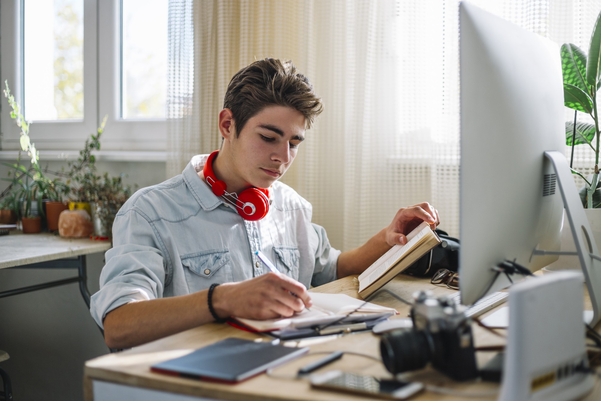A young person with headphones around their neck writes in a notebook at a desk with a computer, camera, and plants in a bright room.