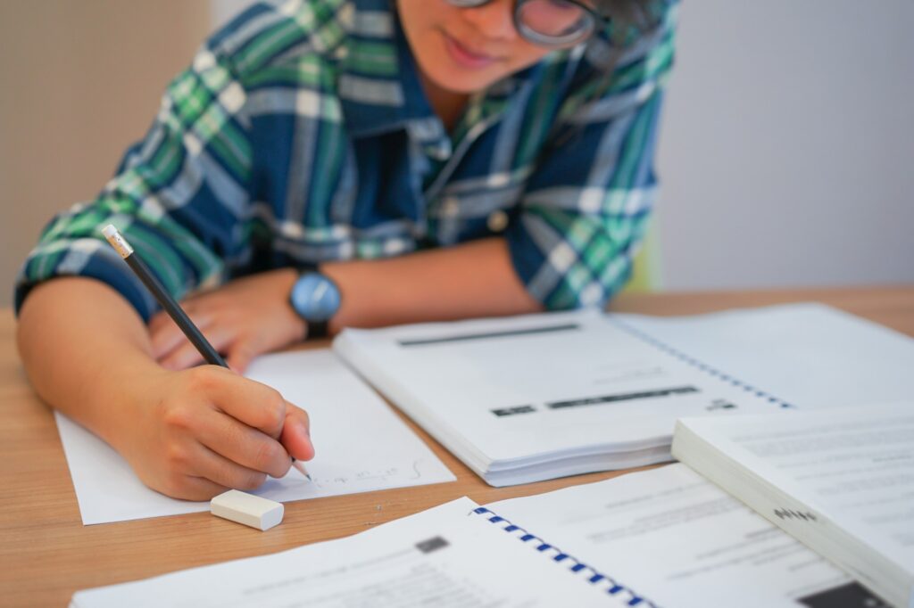 A person wearing glasses and a plaid shirt is writing on paper at a desk covered with notebooks, papers, and an eraser. The focus is on their hand and study materials.