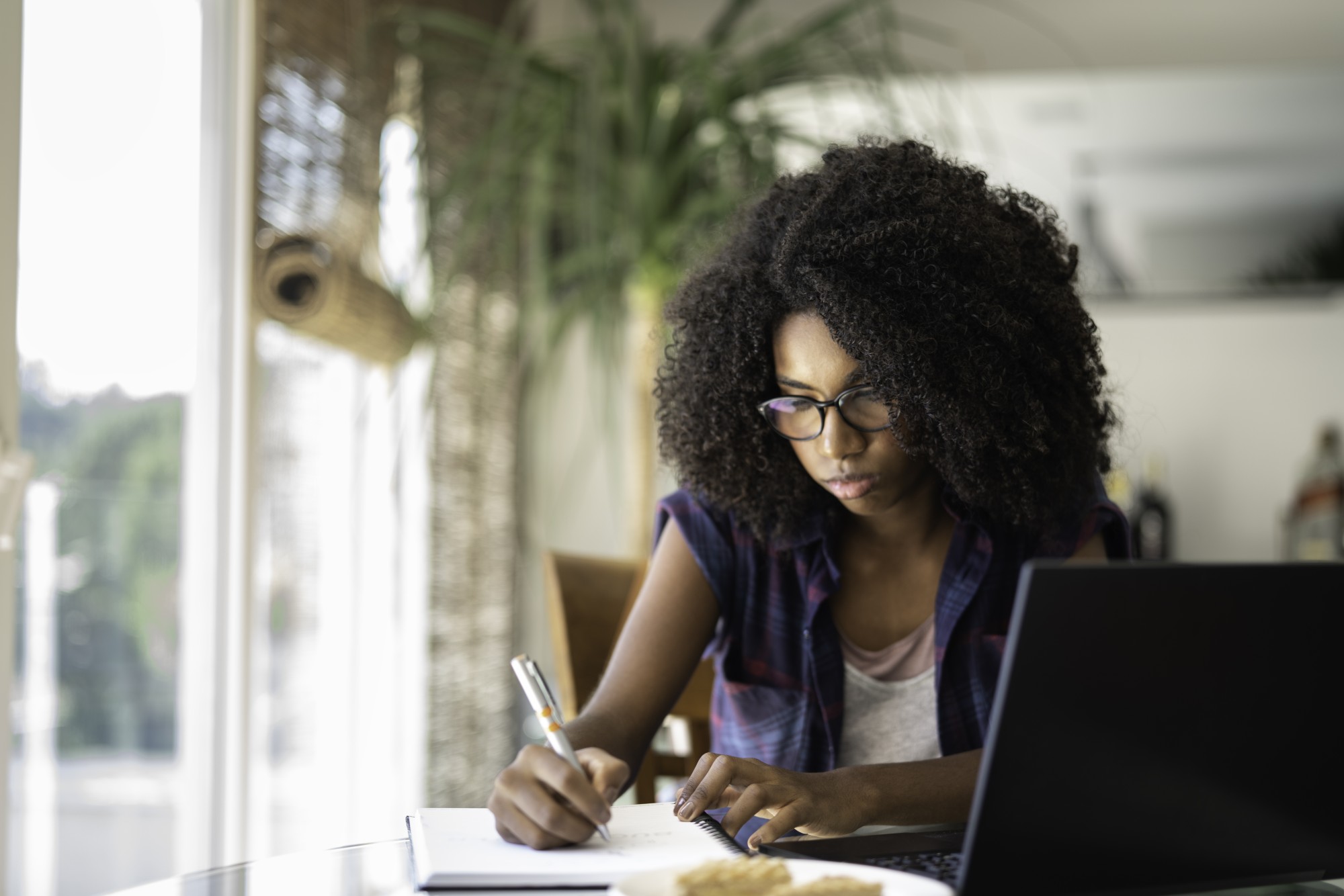 A woman with curly hair and glasses sits at a table, writing in a notebook with a pen, while using a laptop. She appears focused, and there is a blurred plant and window in the background.