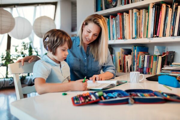 A woman helps a young child with homework at a table filled with colored pencils and art supplies, in a bright room with bookshelves and large paper lanterns.