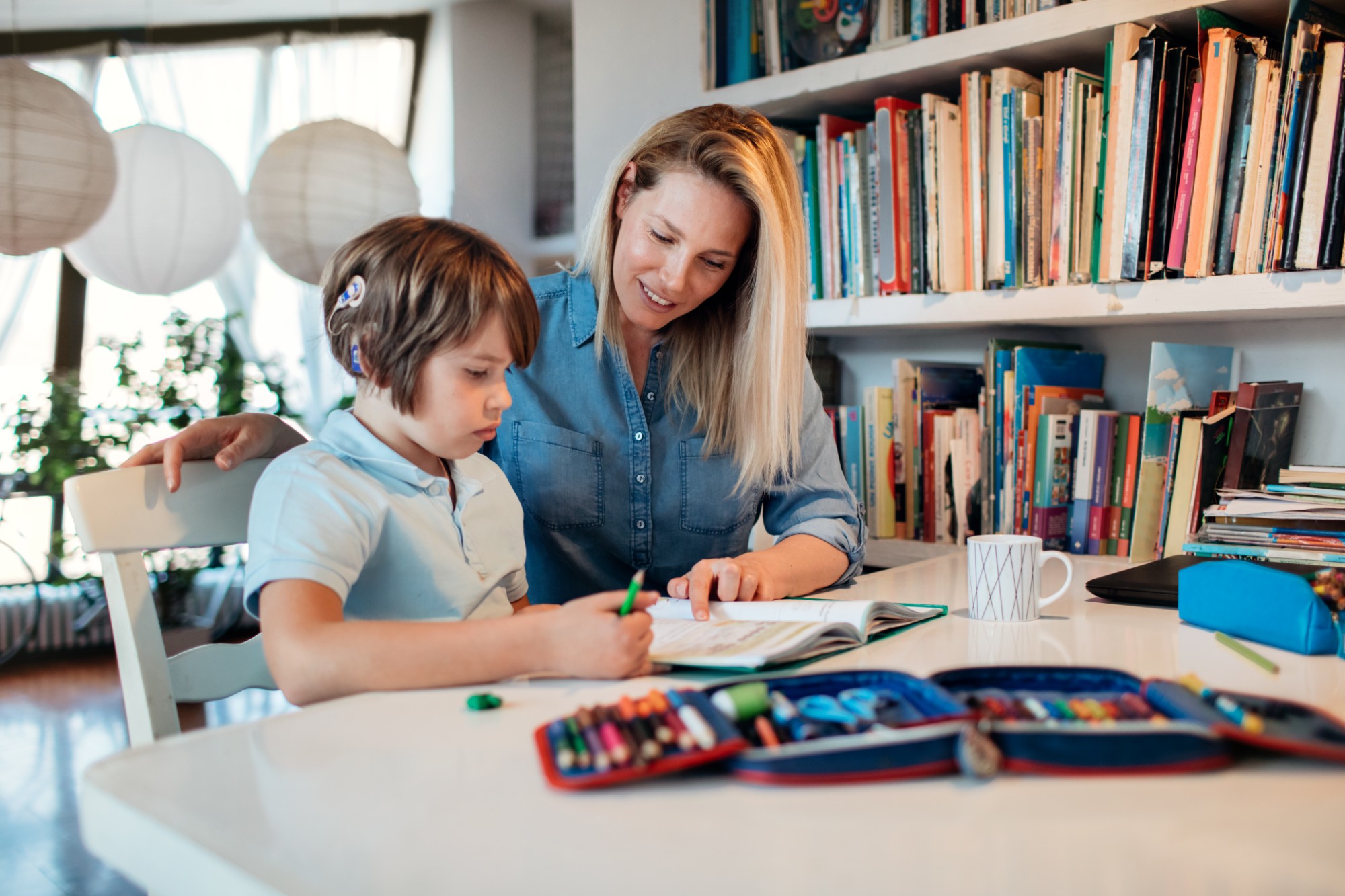 A woman helps a young child with homework at a table filled with colored pencils and art supplies, in a bright room with bookshelves and large paper lanterns.