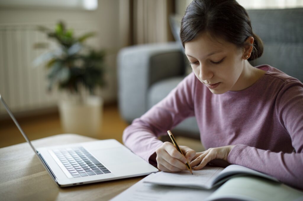 A young person in a pink sweater writes in a notebook while sitting at a table with an open laptop and book, working in a cozy room with a plant and sofa in the background.