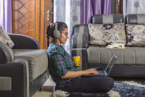 A woman wearing headphones sits on the floor, leaning against a sofa, using a laptop and holding a yellow mug. She is in a living room with patterned sofas, books, and purple curtains.