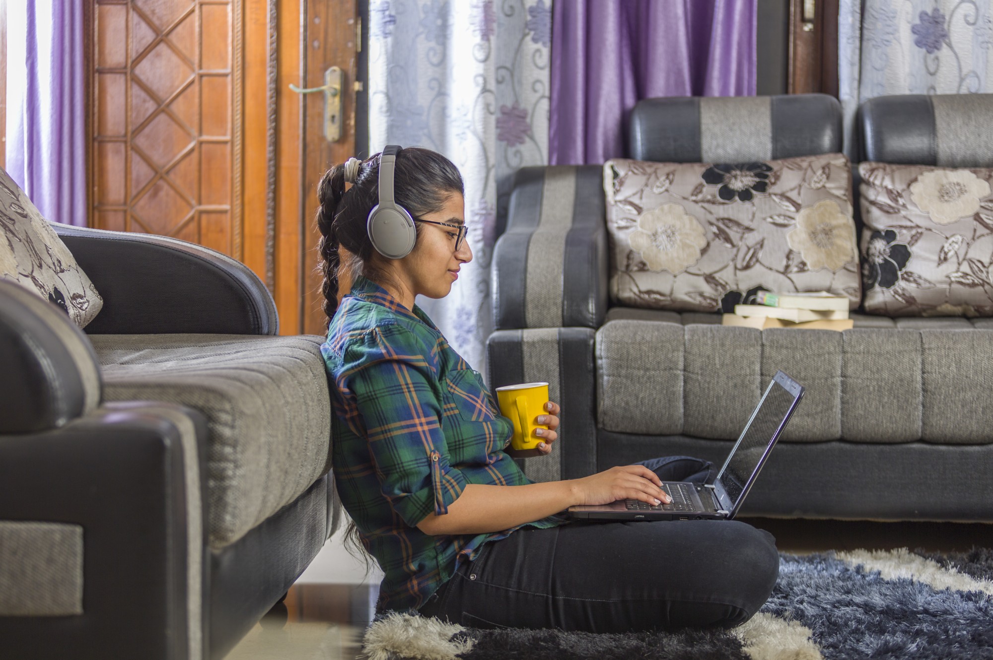 A woman wearing headphones sits on the floor, leaning against a sofa, using a laptop and holding a yellow mug. She is in a living room with patterned sofas, books, and purple curtains.
