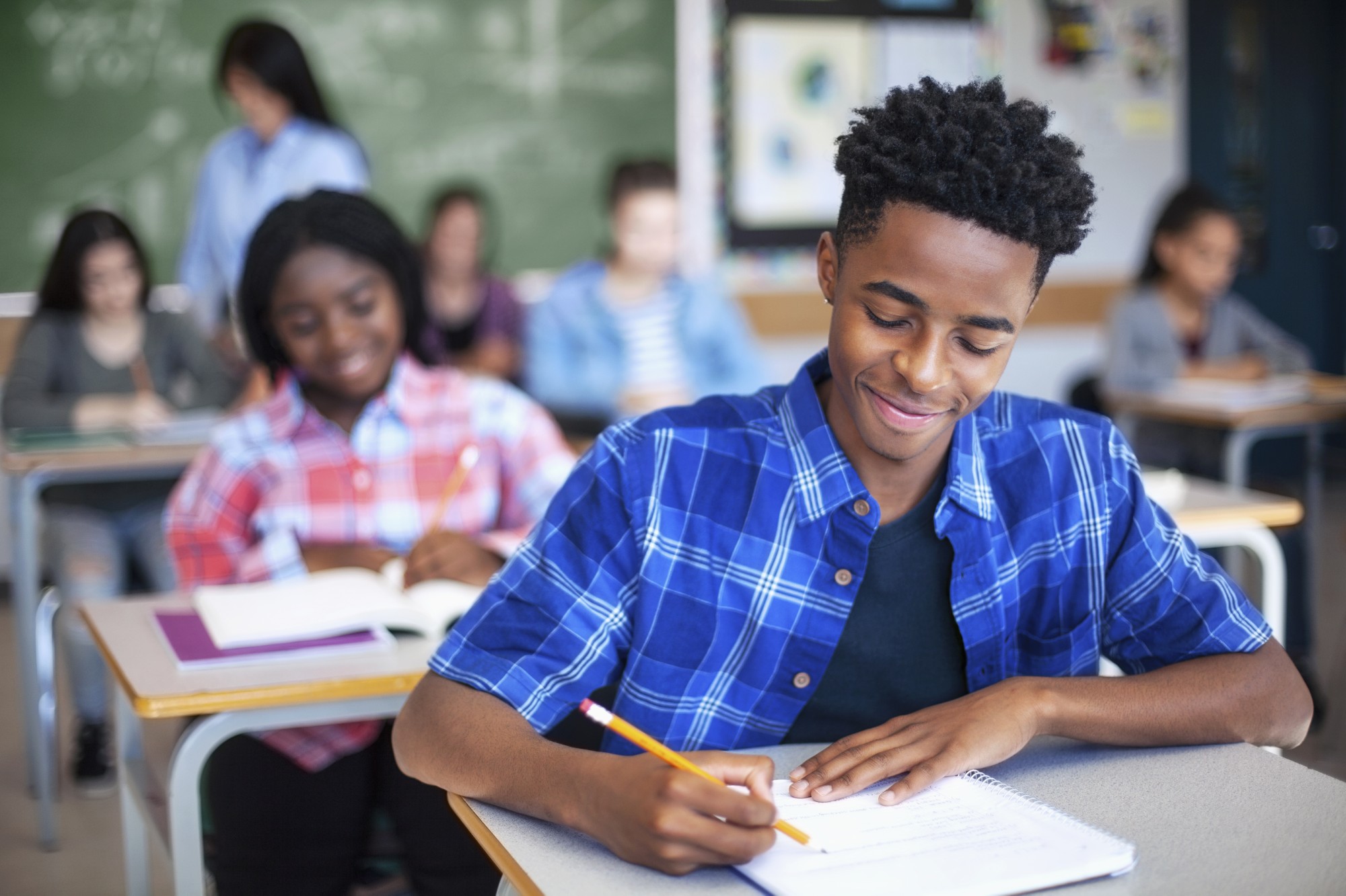 A student in a blue plaid shirt smiles while writing in a notebook at his desk. Other students are seated behind him, and a teacher stands near the chalkboard in a classroom setting.