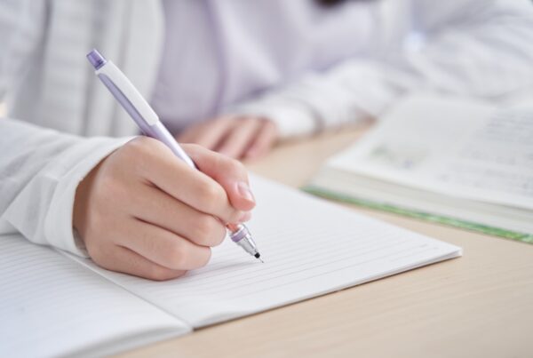 A person writes in a lined notebook with a purple pen, while an open book lies on the table nearby. The setting suggests studying or note-taking.