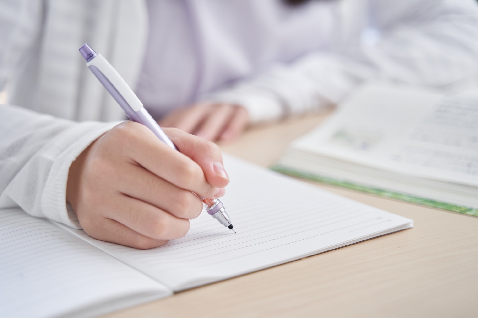 A person writes in a lined notebook with a purple pen, while an open book lies on the table nearby. The setting suggests studying or note-taking.