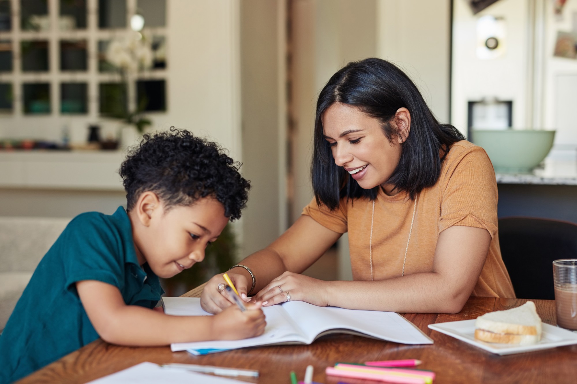 A woman and a young boy sit at a table, smiling and working together on a drawing or homework in a notebook. Colored pencils, paper, and a plate with a sandwich are on the table.