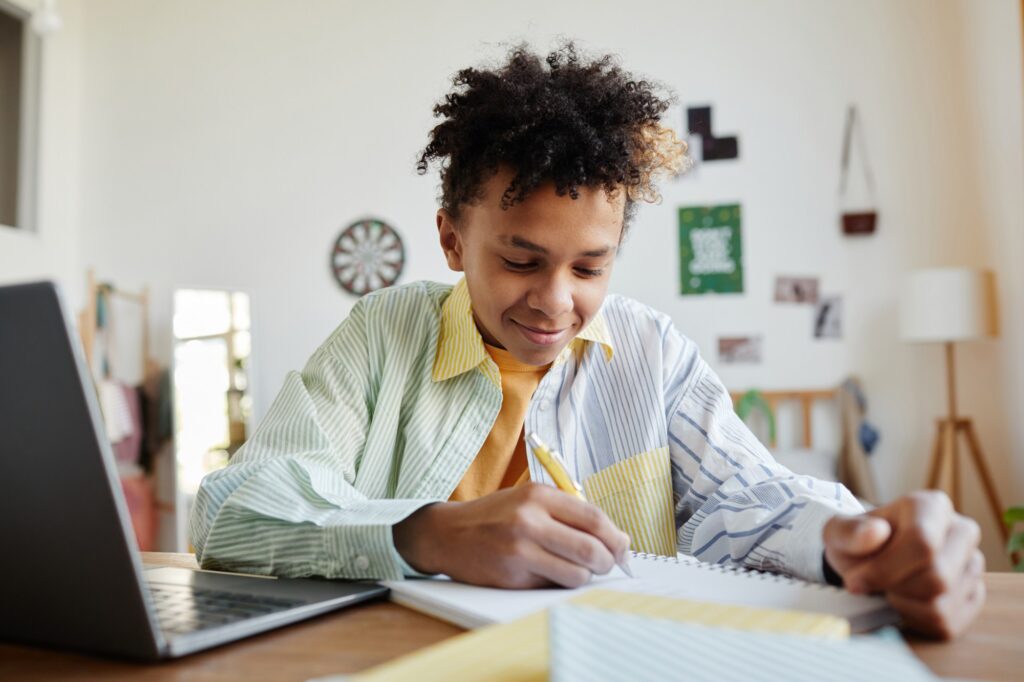 A smiling teenager sits at a desk writing in a notebook, with a laptop open nearby in a brightly lit room decorated with pictures and a dartboard.