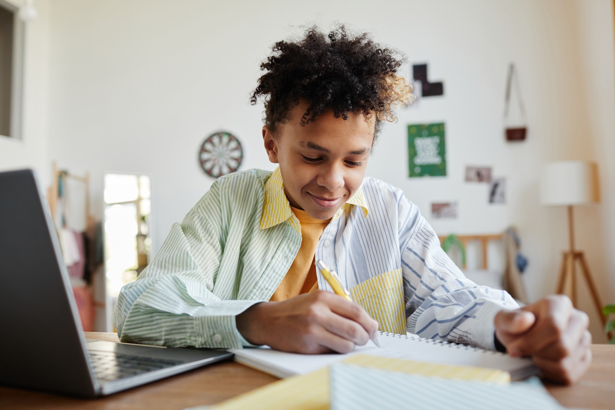 A smiling teenager sits at a desk writing in a notebook, with a laptop open nearby in a brightly lit room decorated with pictures and a dartboard.