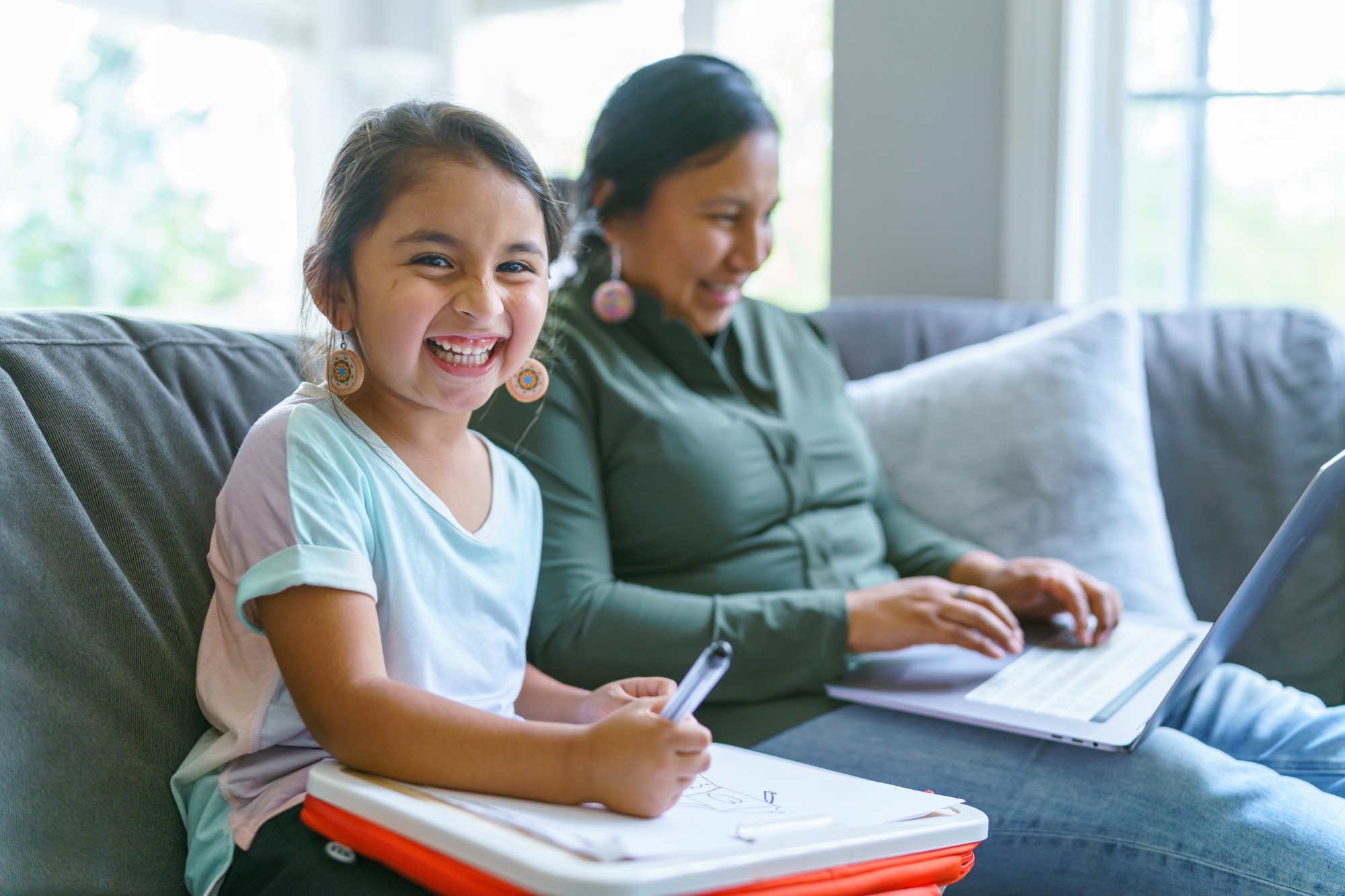 A young girl smiling and holding a marker sits on a couch beside an adult working on a laptop. The room is bright with natural light coming through large windows in the background.