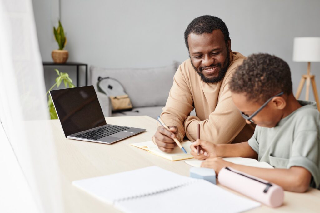 A man smiles while helping a child with homework at a desk. A laptop, notebook, and pencil case are on the table, and they are both writing in notebooks in a bright, cozy room.