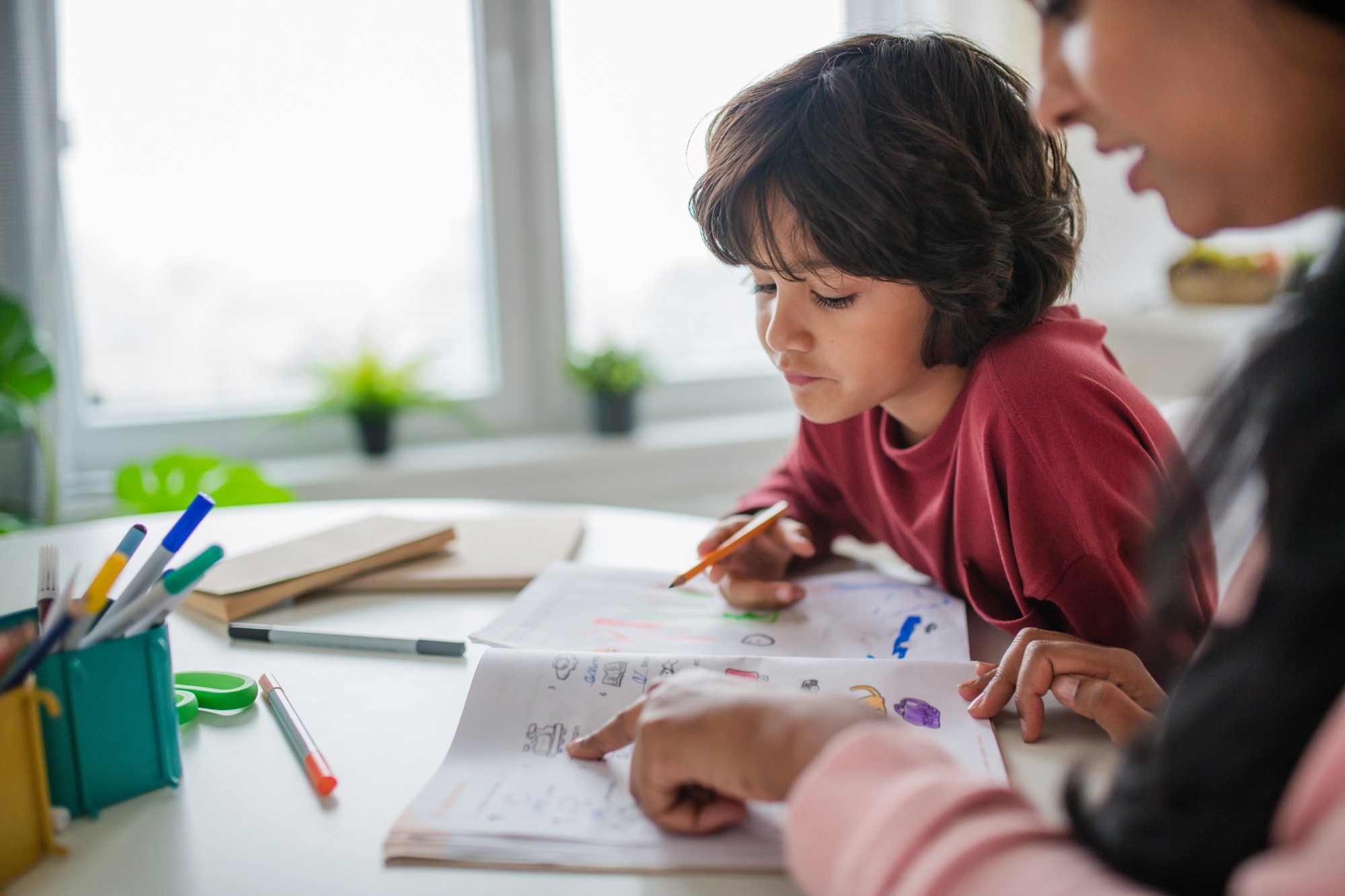 A child sits at a desk doing homework with the help of an adult, who is pointing at a page in a workbook. Colored pencils and school supplies are scattered on the table near a bright window.