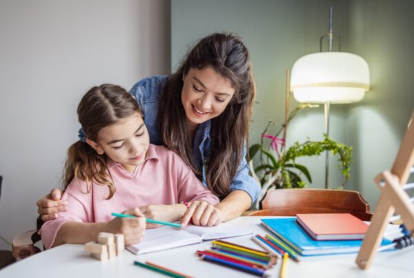 A woman smiles as she helps a young girl with homework at a table covered in colored pencils, books, and notebooks. The girl writes in a notebook while the woman leans over her shoulder, offering support.
