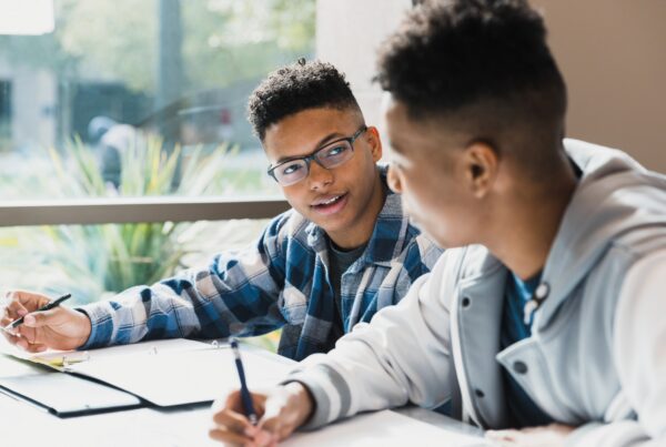 Two teenagers sit at a table with open notebooks, having a conversation and studying together. One wears glasses and a plaid shirt, while the other wears a light-colored jacket. Sunlight comes through a window behind them.