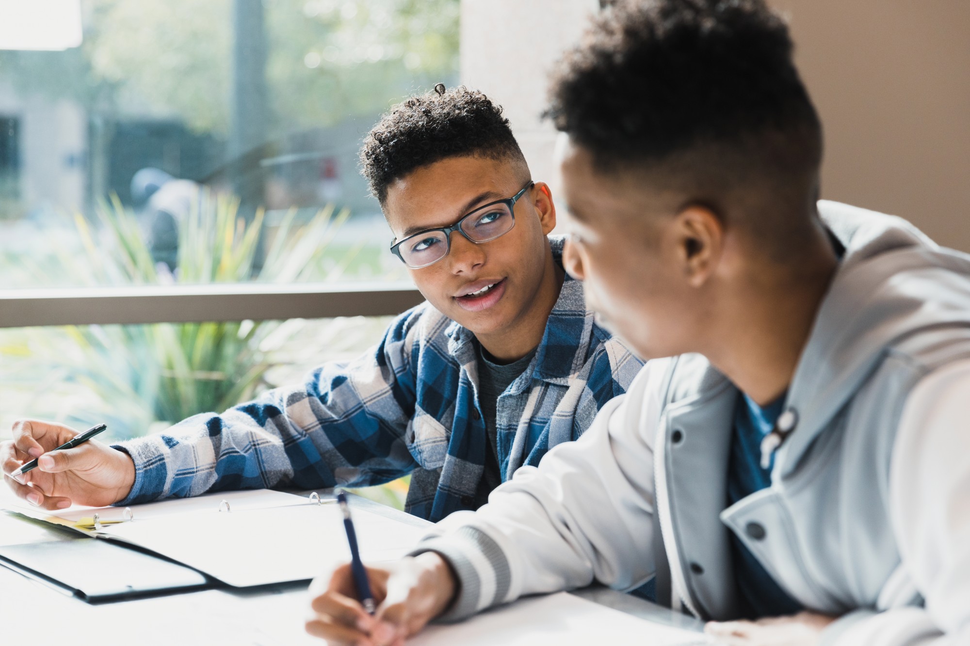 Two teenagers sit at a table with open notebooks, having a conversation and studying together. One wears glasses and a plaid shirt, while the other wears a light-colored jacket. Sunlight comes through a window behind them.