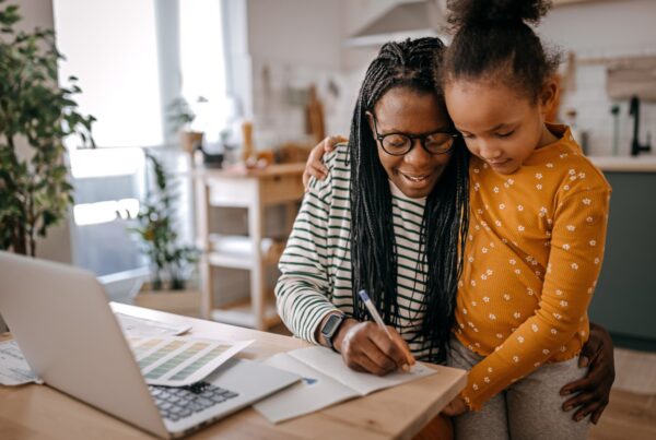 A woman with long braids and glasses writes on paper at a kitchen table with a laptop, while a young girl in a yellow polka-dot shirt hugs her and looks on. They both appear happy and engaged.
