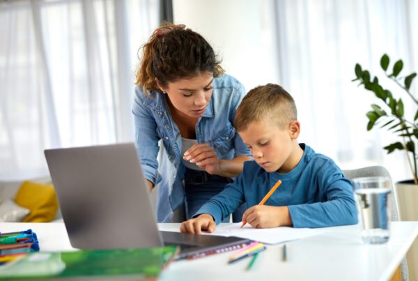 A woman leans over to help a young boy who is writing on paper at a table with a laptop, colored pencils, books, and a glass of water in a bright room with large windows and a plant in the background.