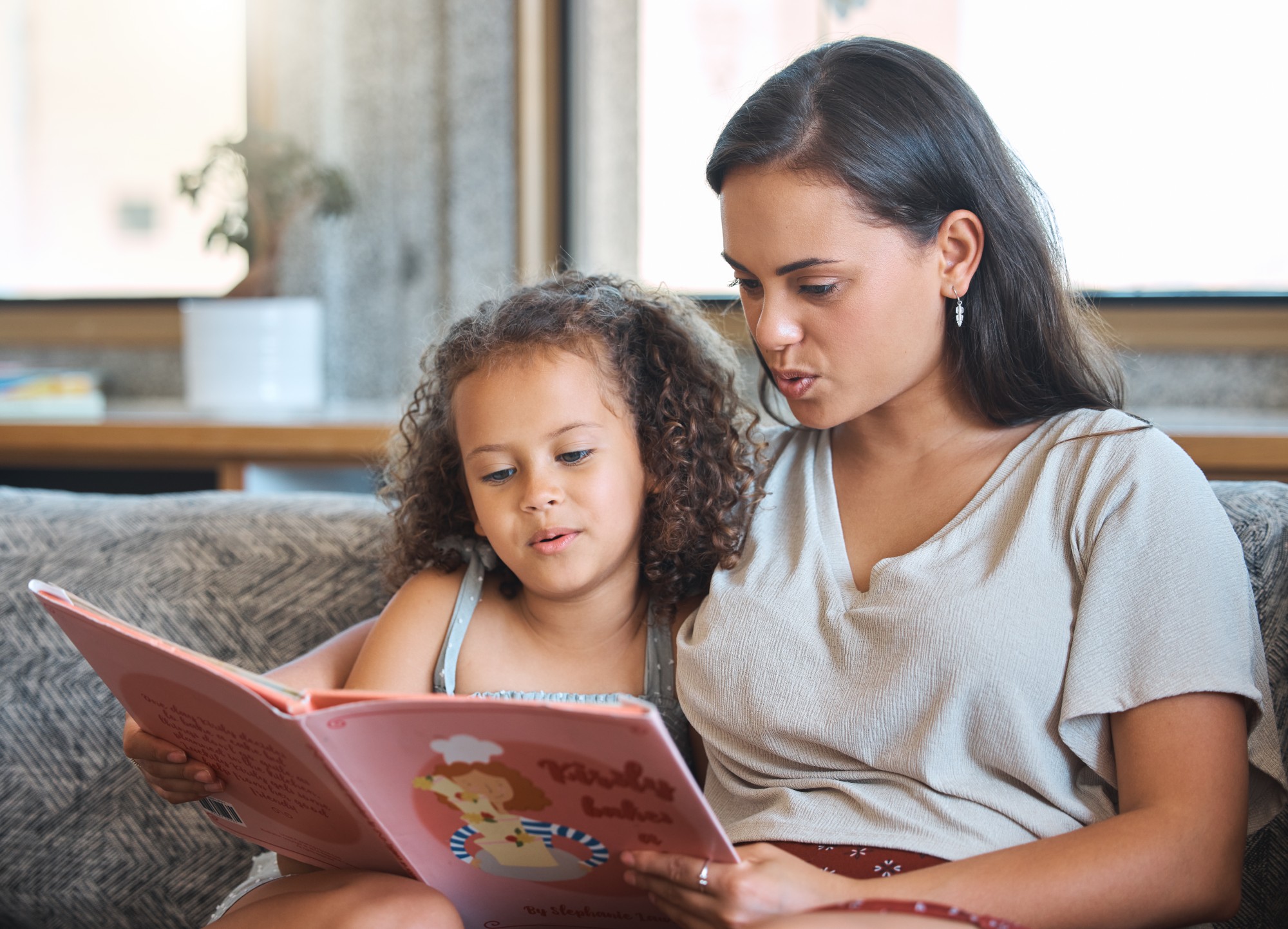 A woman and a young girl sit closely on a couch, reading a colorful children’s book together. Both are engaged with the story, appearing relaxed and focused.