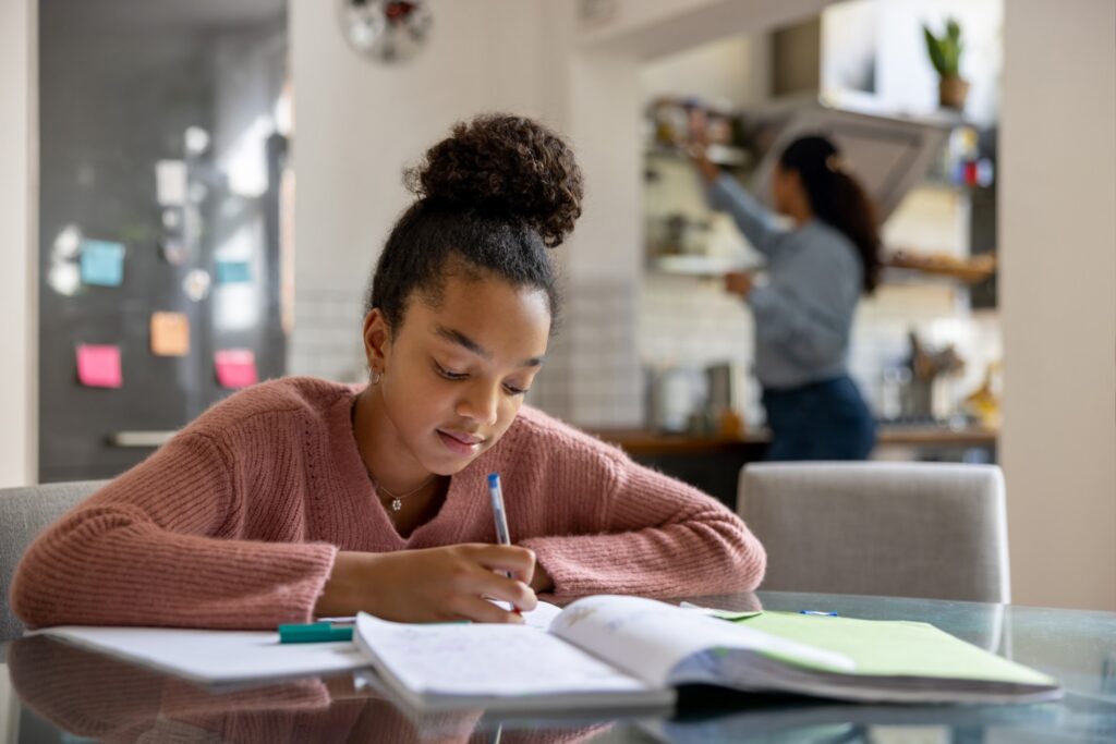 A girl sits at a table doing homework with notebooks and pens, while an adult stands in the kitchen in the background. The room looks bright and organized.