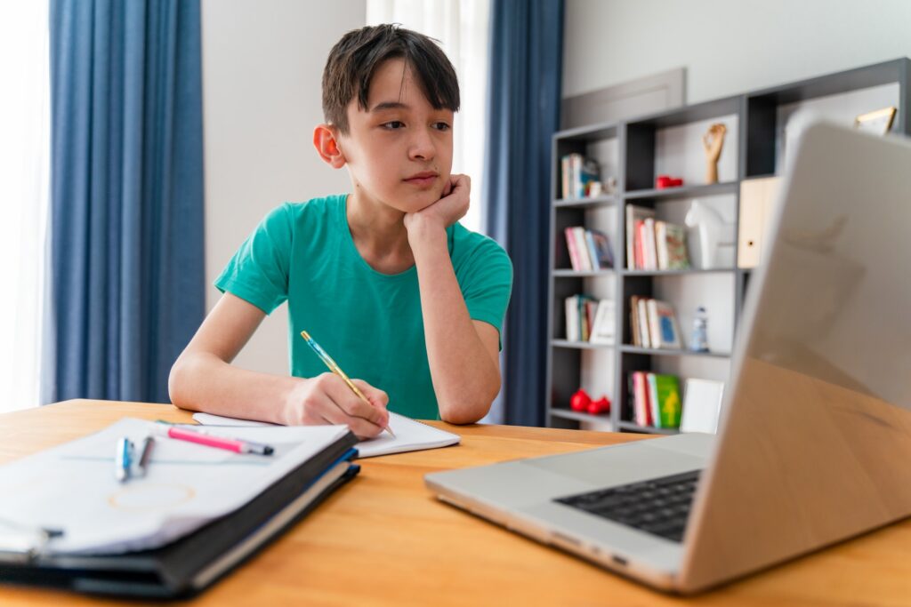 A boy in a teal shirt sits at a desk, writing in a notebook while looking at a laptop. School supplies are on the desk, and a bookshelf is in the background, suggesting he is studying or doing homework.