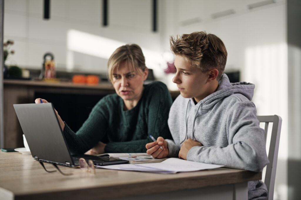 A woman and a teenage boy sit at a kitchen table, looking at a laptop together. The woman gestures at the screen while the boy listens, holding a pencil. Open notebooks and papers are spread out on the table.