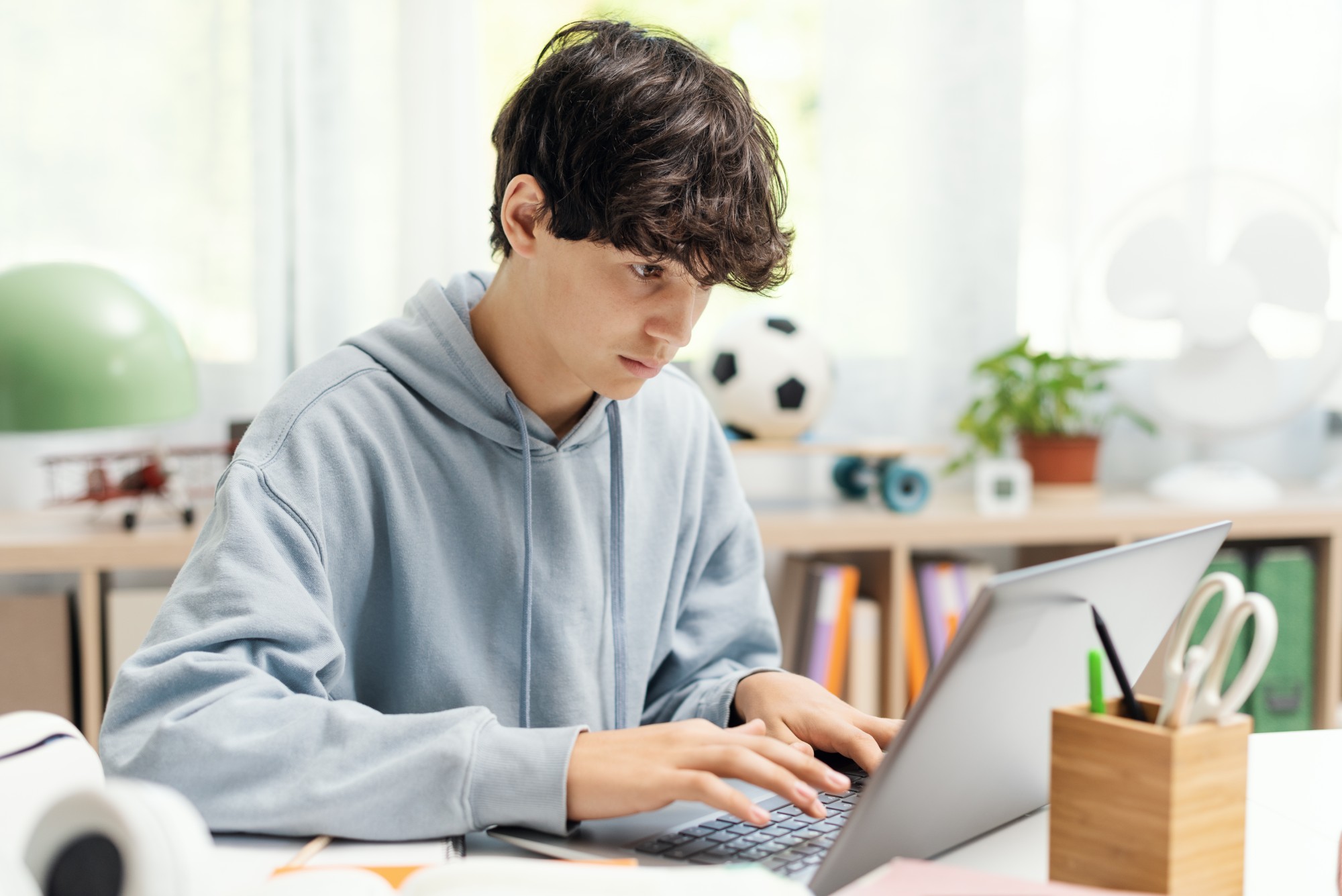 A teenage boy wearing a light blue hoodie sits at a desk typing on a laptop. The background includes books, a plant, a soccer ball, and various stationery items.