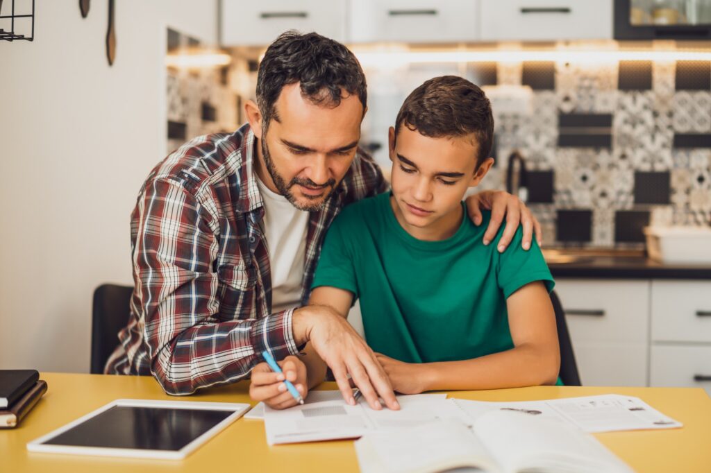 A man helps a boy with homework at a kitchen table, pointing at a notebook. The boy listens attentively. Books and a tablet are on the table, with a modern kitchen in the background.