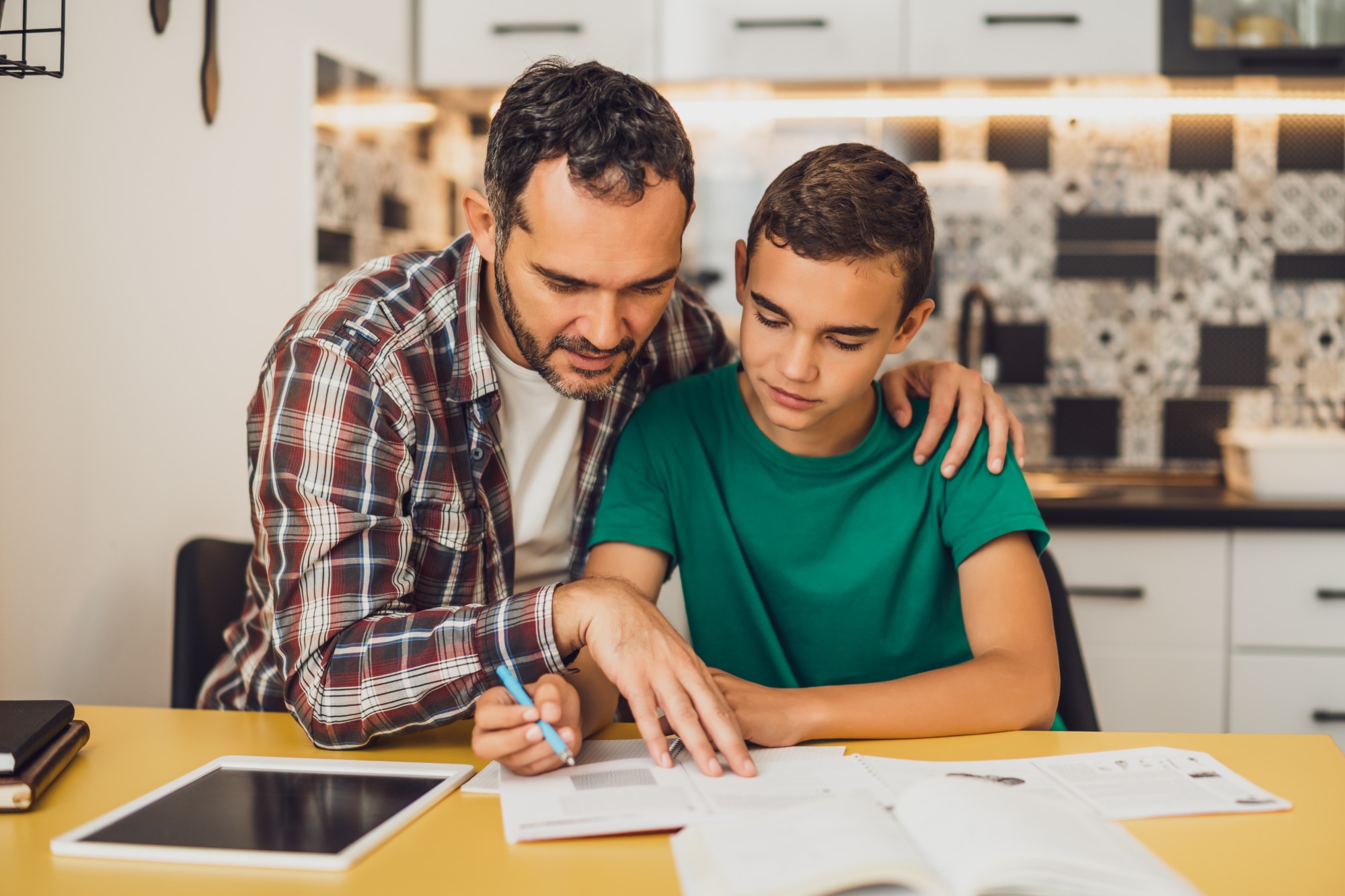 A man helps a boy with homework at a kitchen table, pointing at a notebook. The boy listens attentively. Books and a tablet are on the table, with a modern kitchen in the background.