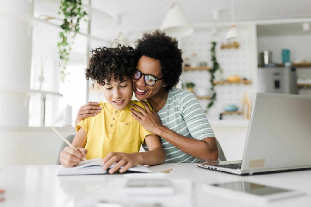 A smiling woman hugs a child who is writing in a notebook at a table with a laptop in a bright, modern kitchen. Both appear happy and engaged in the activity.
