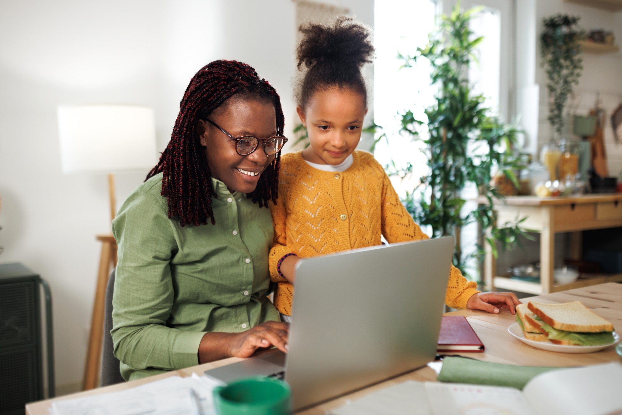 A woman wearing glasses and a green shirt sits at a table using a laptop, smiling, with a young girl in a yellow sweater standing beside her. A plate with a sandwich is on the table. They are in a bright, cozy room.