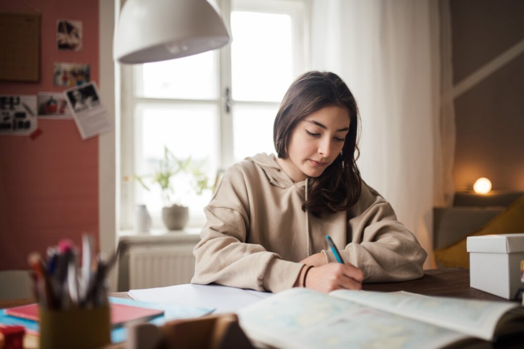 A young woman with long dark hair sits at a desk by a window, writing in a notebook. She is wearing a beige hoodie, and the room appears cozy with soft lighting and various study supplies around her.