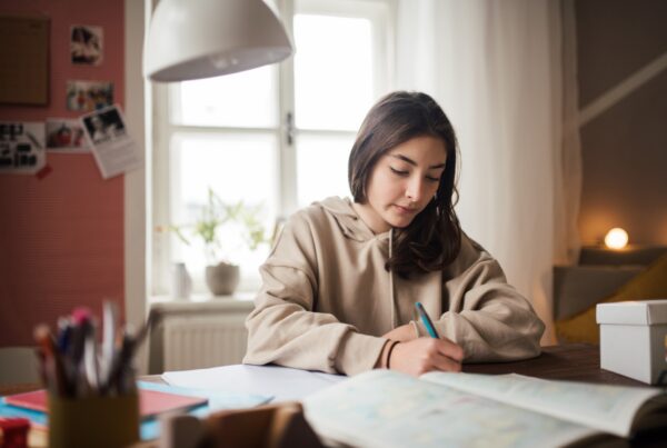 A young woman with long dark hair sits at a desk by a window, writing in a notebook. She is wearing a beige hoodie, and the room appears cozy with soft lighting and various study supplies around her.