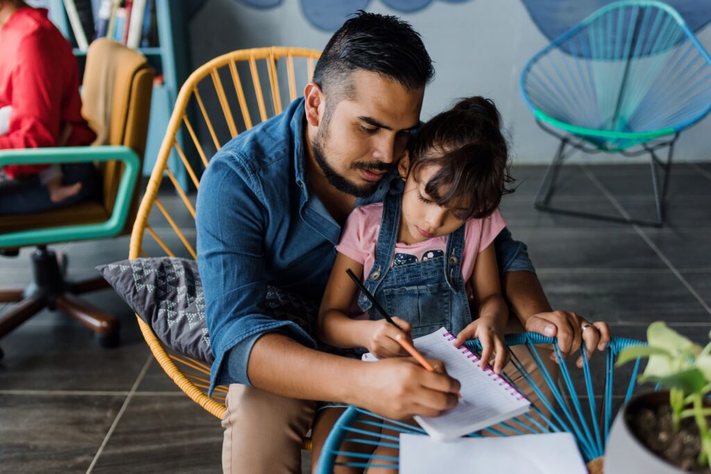 A man sits closely with a young girl on his lap, helping her write in a notebook at a round table. Both appear focused, and the setting is indoors with colorful chairs in the background.