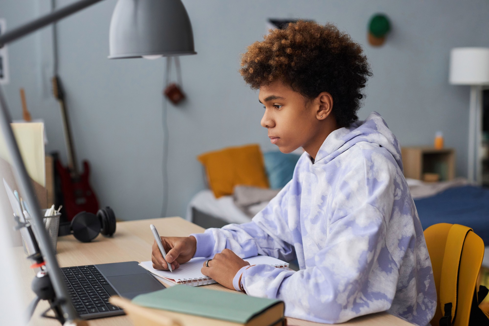A teenage student wearing a tie-dye hoodie sits at a desk, writing in a notebook while looking at a laptop screen in a bedroom. The room has a bed, a guitar, and books visible in the background.
