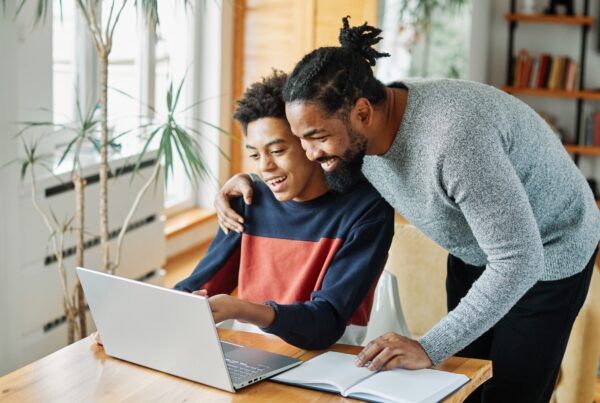 A smiling adult and a teenager sit together at a table, looking at a laptop screen. The adult has one arm around the teens shoulder. An open notebook lies nearby in a bright, cozy room with plants and bookshelves.