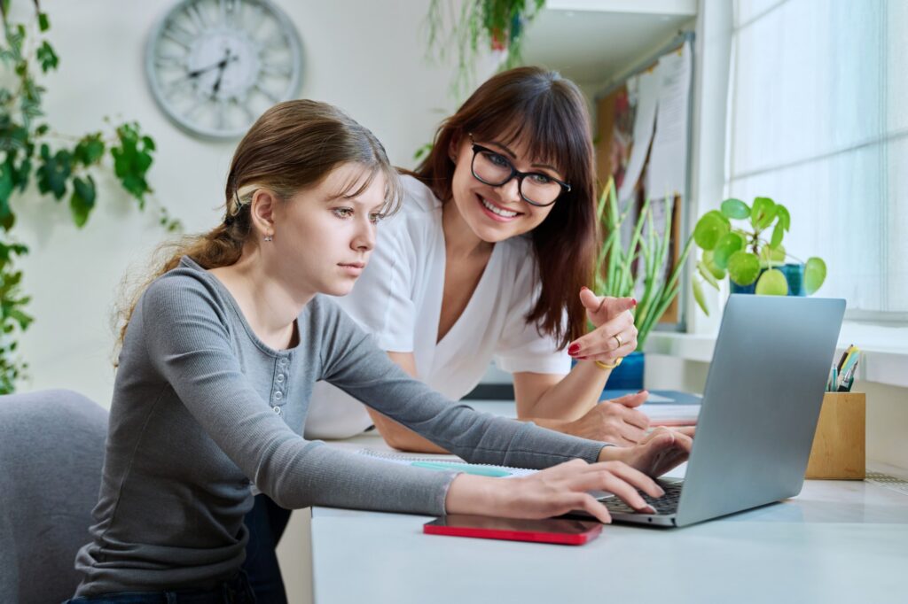 A teenage girl types on a laptop at a desk while an adult woman beside her, smiling and wearing glasses, points at the screen. The room has plants, a clock, and sunlight coming through a window.