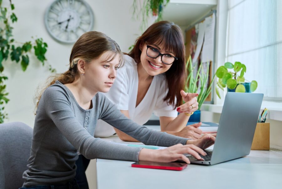 A teenage girl types on a laptop at a desk while an adult woman beside her, smiling and wearing glasses, points at the screen. The room has plants, a clock, and sunlight coming through a window.