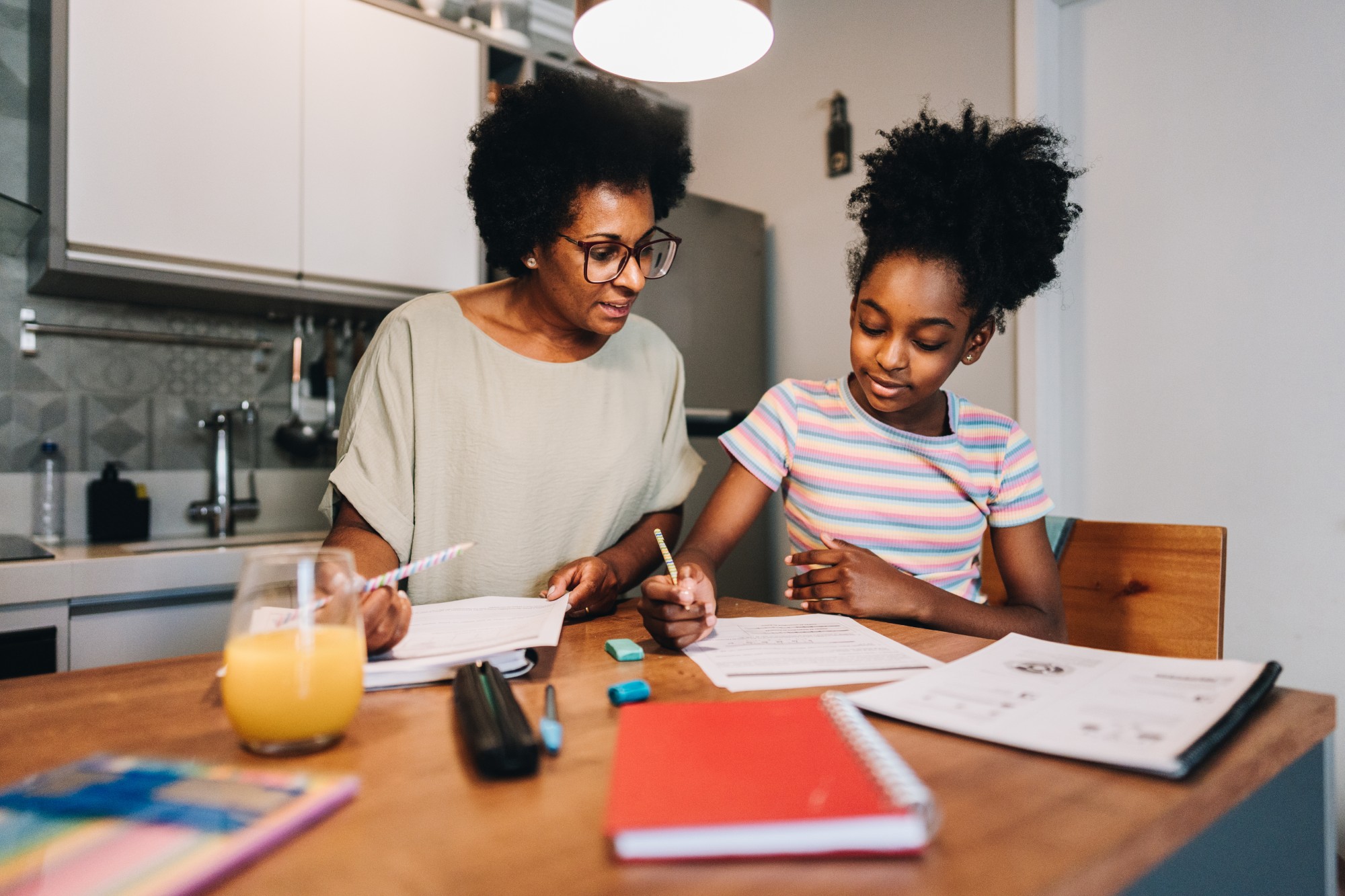 A woman and a young girl sit at a kitchen table working on homework together. The table has notebooks, papers, pens, erasers, and a glass of orange juice on it. Both are focused and writing.