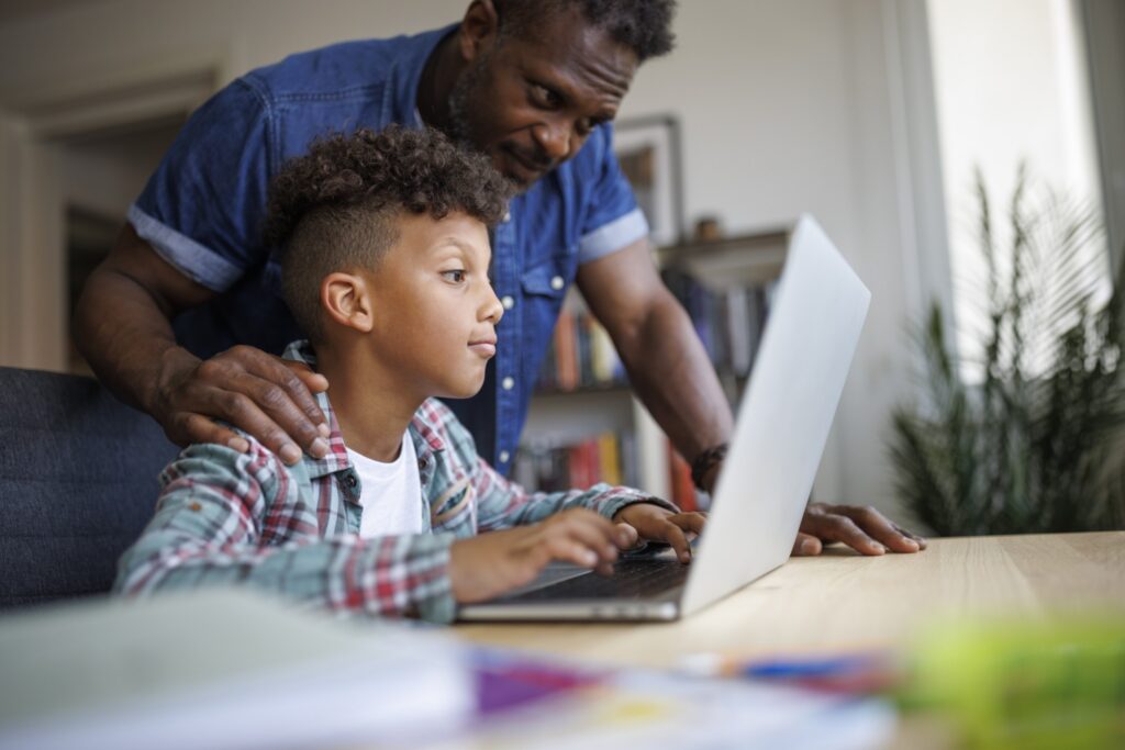 A man stands behind a young boy who is focused on a laptop, gently resting his hands on the boys shoulders. They are indoors, and bookshelves and plants are visible in the background.