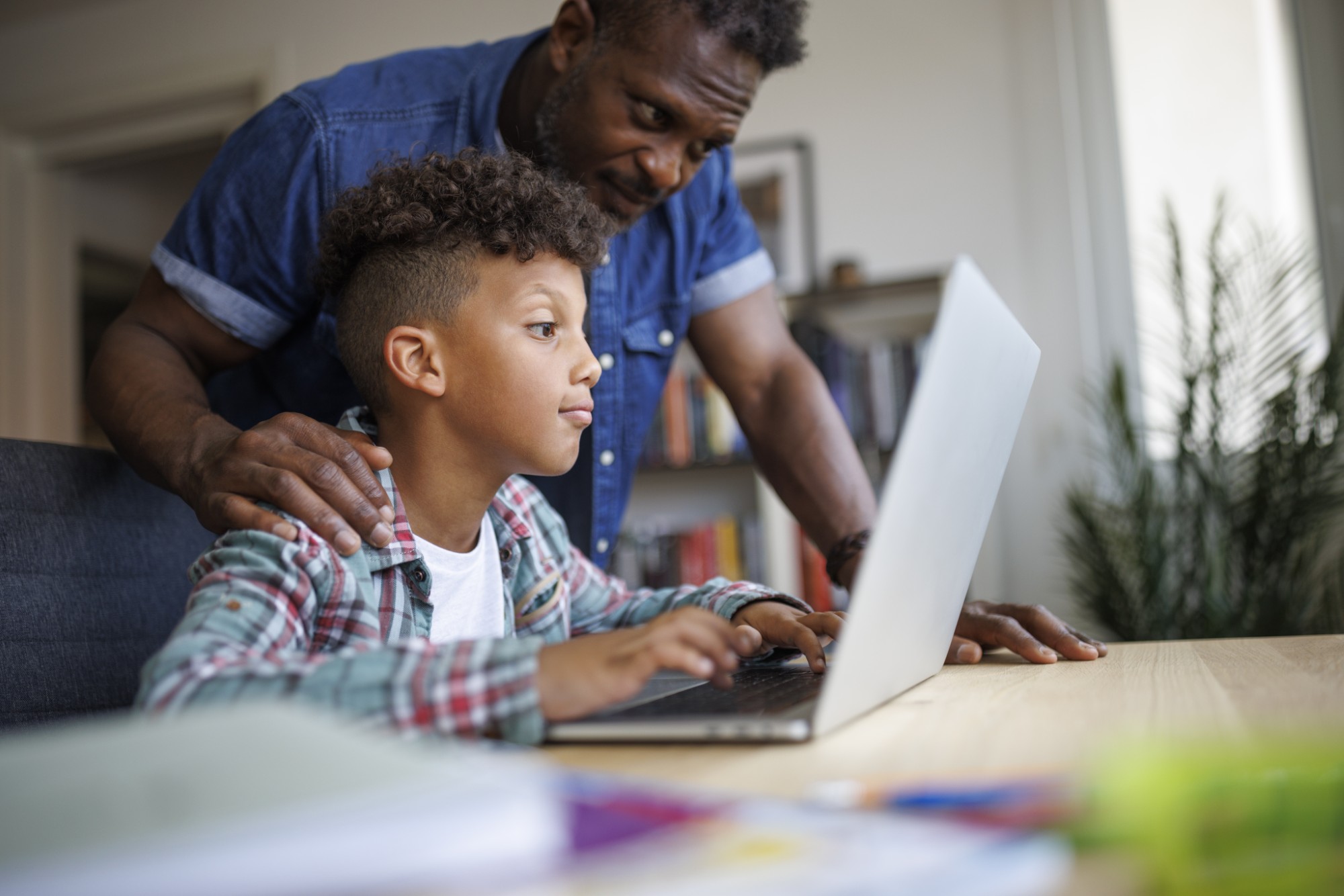 A man stands behind a young boy who is focused on a laptop, gently resting his hands on the boys shoulders. They are indoors, and bookshelves and plants are visible in the background.