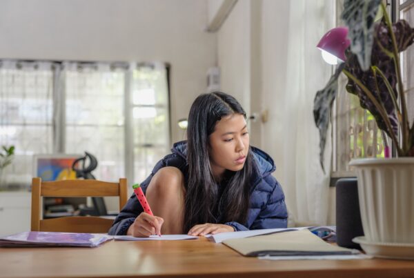 A young woman with long dark hair sits at a table, writing in a notebook with a pink highlighter. She is wearing a dark jacket and is surrounded by books, a potted plant, and a lamp in a well-lit room.