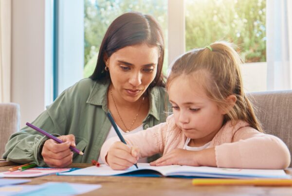 A woman and a young girl sit at a table together, both holding colored pencils and drawing in a notebook, with sunlight streaming through a window behind them.