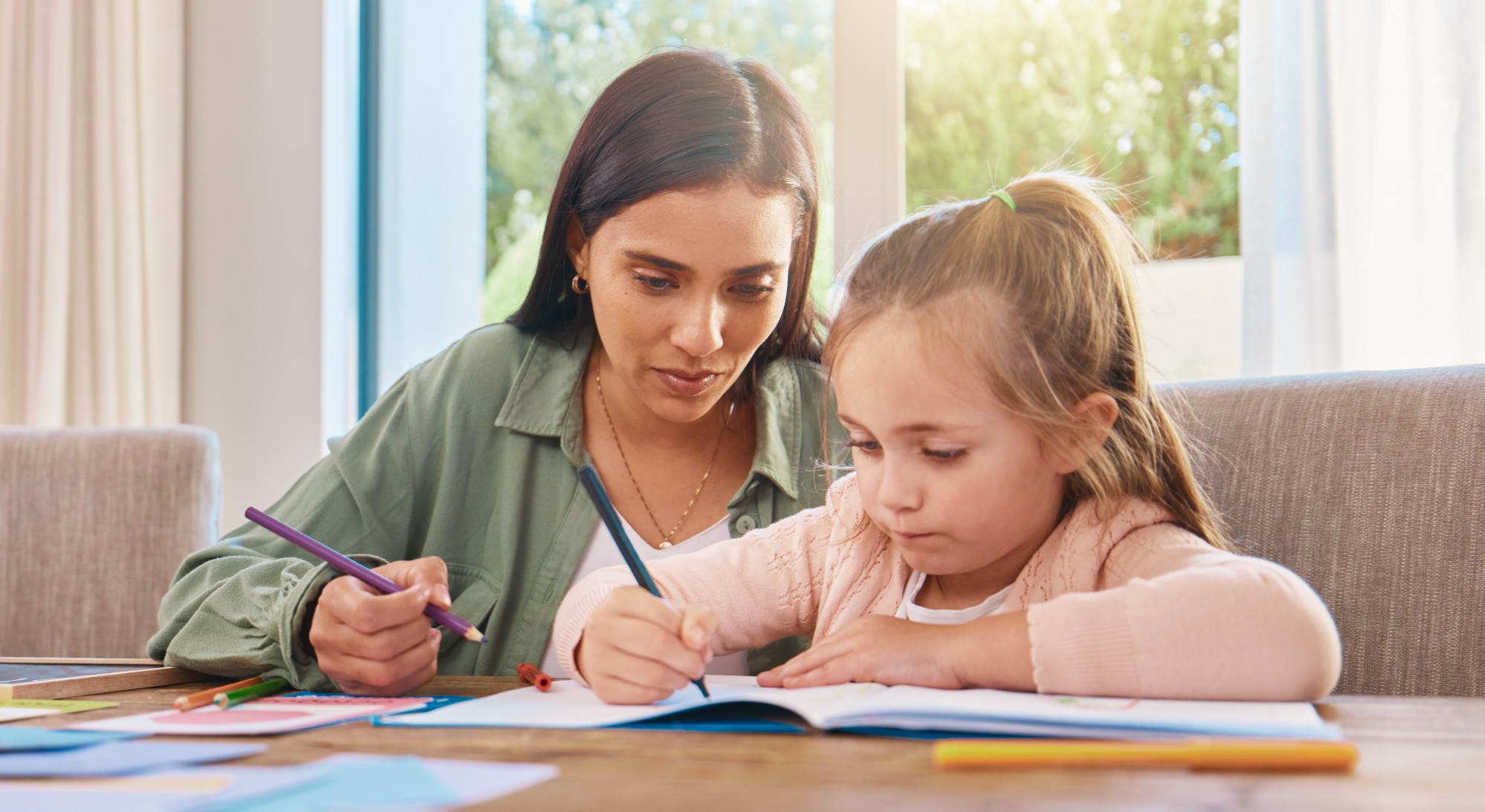 A woman and a young girl sit at a table together, both holding colored pencils and drawing in a notebook, with sunlight streaming through a window behind them.