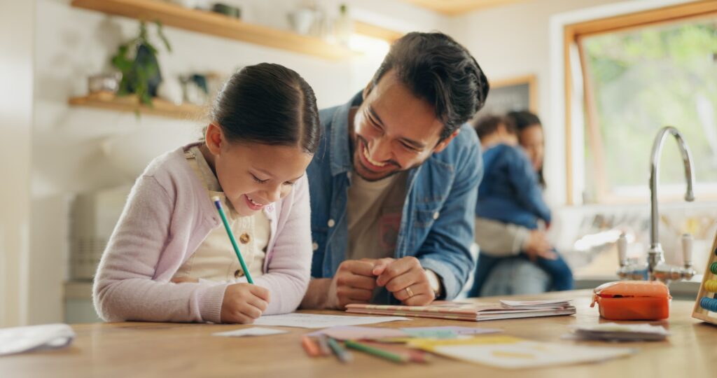 A smiling man helps a young girl with her homework at a kitchen table, while another adult holds a child in the background. The scene is warm and bright, with papers and colored pencils scattered on the table.