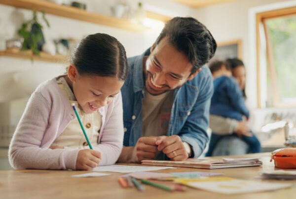 A smiling man helps a young girl with her homework at a kitchen table, while another adult holds a child in the background. The scene is warm and bright, with papers and colored pencils scattered on the table.