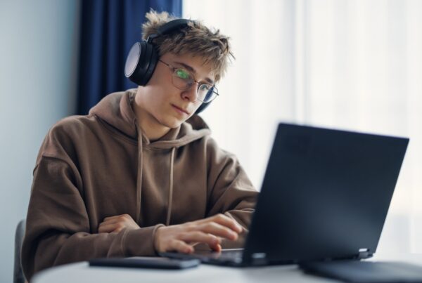 A young person wearing glasses, headphones, and a brown hoodie sits at a desk, focused on using a laptop. Natural light comes from a window in the background.