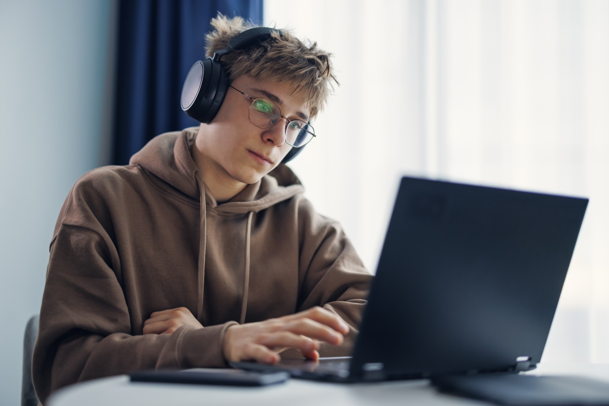 A young person wearing glasses, headphones, and a brown hoodie sits at a desk, focused on using a laptop. Natural light comes from a window in the background.