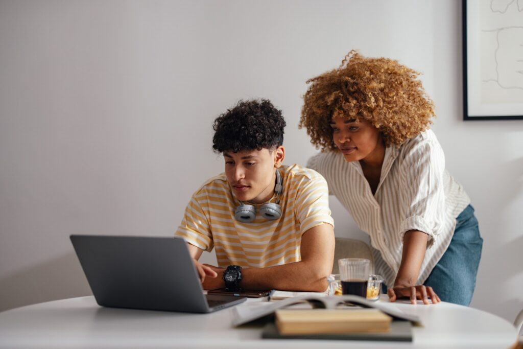 Two young people review something on a laptop at a desk. One sits and types, wearing headphones around his neck; the other stands next to him, leaning over to look at the screen. Books and drinks are on the table.